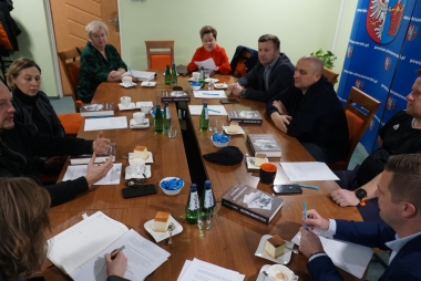 Przy owalnym stole siedzą kobiety i mężczyźni. Przed nimi pisma
