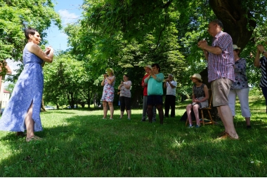 Zajęcia ruchowe w plenerze, prowadzi kobieta w zwiewnej sukni. Przed nią kobiety i mężczyźni 