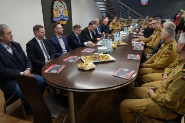 Osoby w garniturach i mundurach przy dużym, owalnym stole. Na stole publikacja, butelki z wodą, kanapki. 