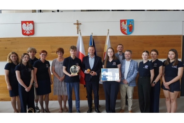 Zdjęcie trzynastu osób w różnym wieku i różnej płci. Kilka osób trzyma statuetki i dyplomy. W tle znajdują się trzy flagi, dwa herby oraz krzyż.