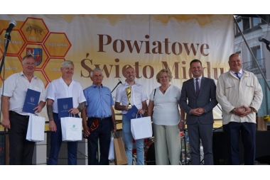 Zdjęcie 6 mężczyzn i 1 kobiety. 4 z Panów trzyma dyplomy, puchary i paczki z prezentami. W tyle baner z napisem Powiatowe Święto Miodu.