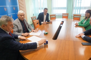 Przy stole siedzi czterech mężczyzn w garniturach i kobieta w zielonej garsonce 
