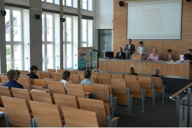 Zdjęcie kilku uczniów siedzących na sali wykładowej i patrzącej w stole podium, na którym siedzi i stoi kilak dorosłych osób. 