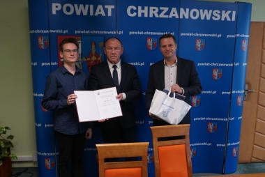 Trzech mężczyzn na tle niebieskiej ścianki z białymi napisami i herbem w kolorze biało, czerwono, żółtym