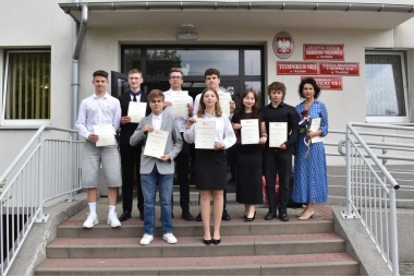 NA ZDJĘCIU 9 OSÓB, UCZNIOWIE ZSTU WRAZ Z OPIEKUNEM W NIEBIESKIEJ SUKIENCE  STOJĄ PRZED SZKOŁĄ  NA SCHODACH, UBRANI W GALOWE STROJE ORAZ PREZENTUJĄ DO ZDJĘCIA DYPLOMY 