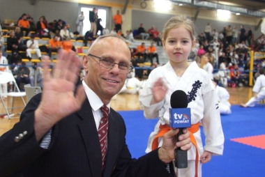 Uśmiechnięty mężczyzna macha ręką do kamery. W drugiej ręce trzyma mikrofon. Obok mała dziewczynka - zawodniczka karate 