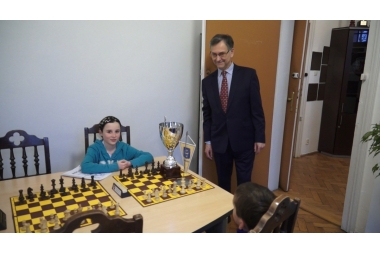 na zdjęciu dziewczynka siedząca przed szachownica, na stoliku złoty puchar obok stoi i spogląda na nią Janusz Szczęśniak