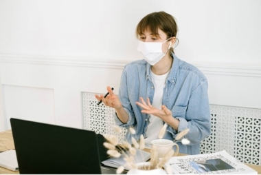 na zdjeciu kobieta w jeansowej koszuli i masce chirurgicznej, siedząca przed laptopem, w dłoni trzyma długospis, na biurku suszone kwiaty oraz ksiazka 