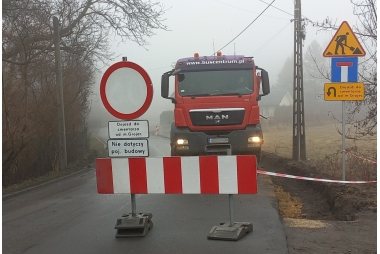 Zdjęcie drogi na której stoją znaki komunikujące o zamknięciu drogi oraz objeździe. Za znakami stoi ciężarówka używana do remontu drogi.