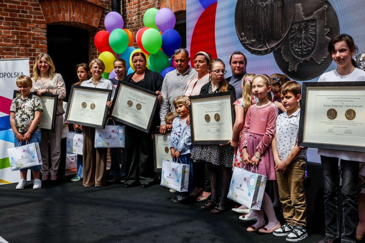 Zdjęcie kilkunastu osób pozujących do zdjęcia. Na zdjęciu są osoby dorosłe oraz dzieci. Większośc z nich trzyma duże dyplomy oraz torby prezentowe. 