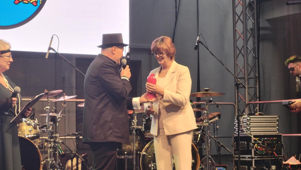 Zdjęcie dwóch osób na scenie. Mężczyzna w cylindrze przekazuje kobiecie statuetkę czerwonego sokoła. W tle instrumenty muzyczne. 