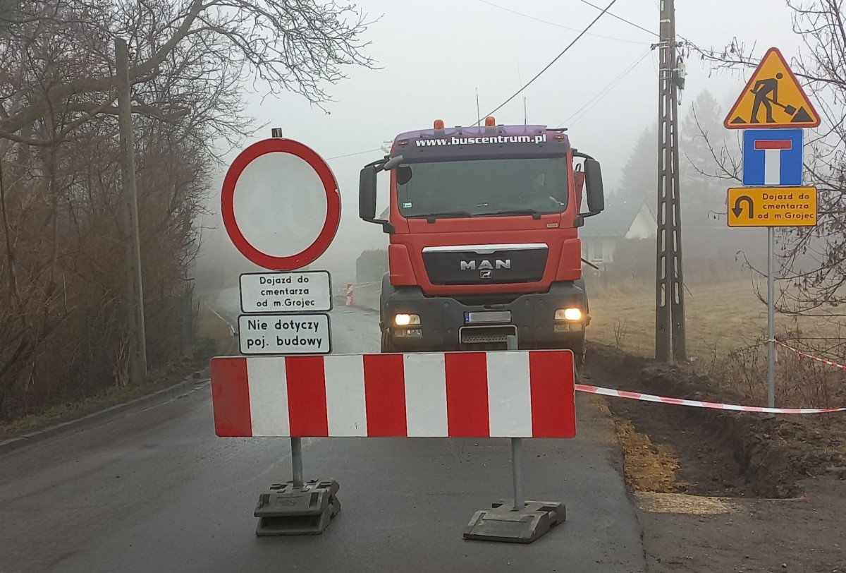 Zdjęcie drogi na której stoją znaki komunikujące o zamknięciu drogi oraz objeździe. Za znakami stoi ciężarówka używana do remontu drogi.